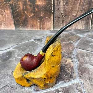 May include: A burgundy smoking pipe with a black stem and gold accent rests on a uniquely shaped, yellow-toned wooden stand. The pipe's bowl has a textured finish. The background features a wood-grain pattern.