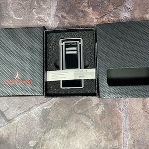 Może przedstawiać: Czarna i srebrna zapalniczka Lotus w otwartym czarnym pudełku. Zapalniczka ma na sobie nazwę marki. Pudełko jest czarne z teksturowanym wzorem. Zapalniczka jest prezentowana na czarnej wkładce z pianki.