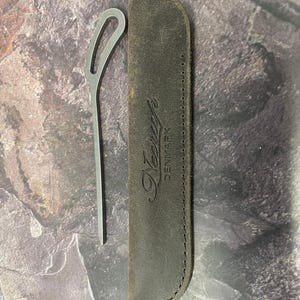 May include: A silver metal tool with a looped handle and a dark brown leather sheath. The sheath is embossed with the word "Neerup DENMARK". The tool and sheath are displayed on a textured surface.