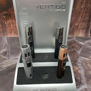 May include: A display stand with four Vertigo by Lotus lighters in silver, black, and brown. The stand is silver with the brand name on the front and back. The lighters are arranged on a black foam base.