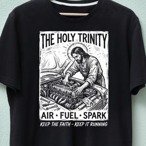 May include: Black t-shirt featuring a graphic of Jesus working on a car engine. The image includes the text "THE HOLY TRINITY" above the image, and "AIR • FUEL • SPARK" and "KEEP THE FAITH - KEEP IT RUNNING" below the image.