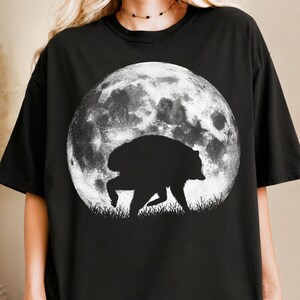 May include: A black short-sleeved t-shirt featuring a large, circular graphic print on the front. The design displays a detailed, textured white full moon in the background with the black silhouette of a bear walking through grass in the foreground.