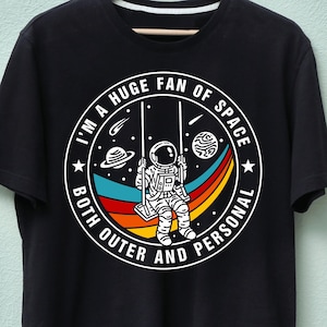 以下が含まれることがあります： 黒いTシャツ。円形のデザインで、ブランコに乗った宇宙飛行士、虹、宇宙の要素が描かれています。「I'M A HUGE FAN OF SPACE BOTH OUTER AND PERSONAL」というテキストが入っています。