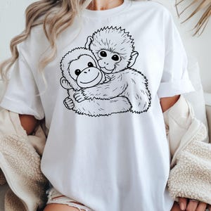 May include: White t-shirt featuring a black and white illustration of two monkeys hugging. The design shows one monkey embracing the other, with detailed line art creating a playful and heartwarming image. The shirt is made of a soft, comfortable material.