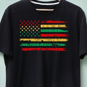 Może przedstawiać: Czarny t-shirt z postarzanym wzorem amerykańskiej flagi, zawierającym czerwone, żółte, zielone i złote paski i gwiazdy. Koszulka jest prezentowana na drewnianym wieszaku na tle jasnozielonej ściany.