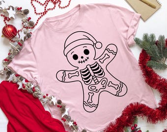 Pode incluir: Camiseta rosa claro com um desenho em preto e branco de um homem de gengibre esqueleto usando um chapéu de Papai Noel. O design inclui costelas, ossos dos braços e pernas. Um design de feriado festivo e único.
