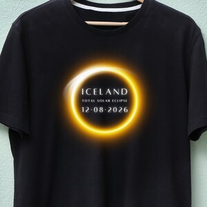 Peut inclure: T-shirt noir avec un motif de cercle lumineux jaune et blanc représentant une éclipse solaire. Le texte "ICELAND TOTAL SOLAR ECLIPSE 12.08.2026" est inclus. Le t-shirt est suspendu à un cintre en bois.