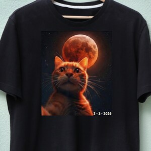 May include: Black t-shirt with a striking image of an orange tabby cat looking up at a large, reddish-orange moon. The date "3-3-2026" is printed below the image. The shirt is made of a soft material.