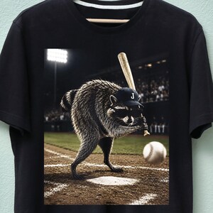 Pode incluir: Uma t-shirt preta com uma estampa gráfica de um guaxinim usando um boné de beisebol com a letra J e óculos de sol, segurando um taco de beisebol de madeira em um campo de beisebol com uma bola voando em sua direção no estádio.