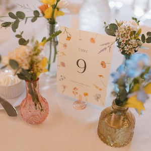 Peut inclure: Une table décorée avec des compositions florales dans de petits vases. Une carte de numéro de table affiche "TABLE 9". Les vases sont en verre rose, doré et transparent. Les fleurs comprennent des fleurs jaunes, blanches et bleues.