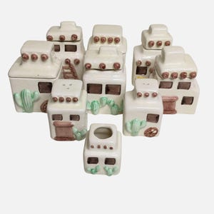 Pode incluir: Uma coleção de saleiros e pimenteiros de cerâmica em estilo sudoeste e um porta-palitos. O conjunto apresenta peças em forma de casa esbranquiçada com detalhes castanhos, detalhes de cactos e pequenos pontos castanhos. O porta-palitos está no centro.