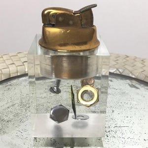 Op de afbeelding: Een vintage goudkleurige aansteker met een getextureerd oppervlak, geplaatst op een helder acrylblok. Het blok bevat diverse hardware-artikelen, waaronder een schroef, een moer en een ring. De aansteker heeft een scharnierend deksel en een ontstekingsmechanisme.