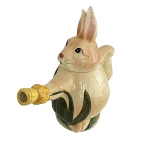 May include: A ceramic teapot in the shape of a bunny rabbit. The teapot is primarily beige with green and yellow accents. The bunny has upright ears, a red nose, and black eyes. The spout is yellow, and the base features green leaves.