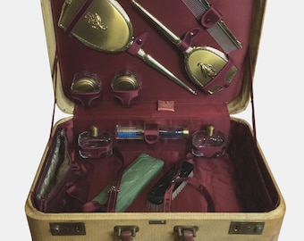 May include: An open vintage travel vanity case with a cream-colored exterior and a burgundy interior. The case contains gold-toned cosmetic accessories, perfume bottles, and grooming tools. The case has a brown handle and gold-colored clasps.