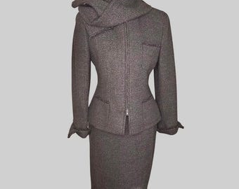 May include: A brown tweed skirt suit with a matching jacket. The jacket has a unique draped collar, a zipper closure, and pockets. The skirt is knee-length. The suit is displayed on a mannequin.
