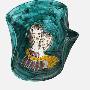 May include: A teal ceramic bowl with a wavy rim, featuring a hand-painted illustration of two embracing figures. The figures have long, light brown hair and are adorned with colorful clothing. The bowl's interior has a textured, painted appearance.