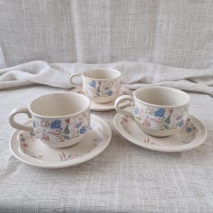May include: Three cream-coloured teacups and saucers with floral patterns. The cups have curved handles and sit on matching saucers. The floral design includes blue, purple, and pink flowers with green leaves. The set is arranged on a neutral-toned fabric.