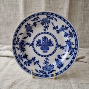 May include: White ceramic plate with a blue floral design. The plate features a central motif of a basket of flowers, surrounded by a circular border and floral patterns. The rim has a detailed blue border.