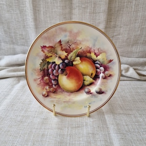 May include: A decorative porcelain plate with a still life of fruit. The design includes ripe peaches, purple grapes, and green leaves. The rim is trimmed with gold, and the plate is displayed on a gold stand.