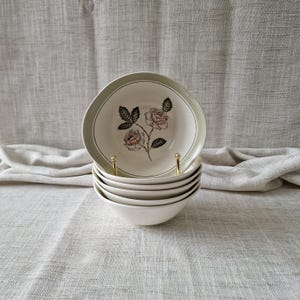 May include: A stack of five off-white ceramic bowls with a light green rim. The top bowl features a detailed illustration of two roses with green leaves and pink-tinted petals centred on the interior base of the bowl.