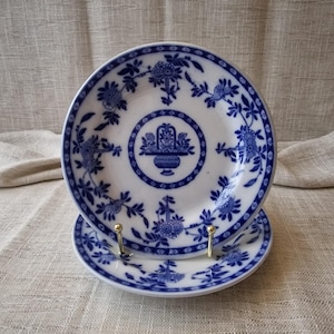 May include: Two white ceramic plates with a blue floral design. The plates feature a central motif of a basket of flowers and are surrounded by a border of floral patterns. The plates are displayed on a gold-coloured stand.