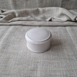 May include: A small, white ceramic trinket box with a round, domed lid. The lid and base are connected by a decorative, rope-like trim. The box sits on a textured, light grey surface.