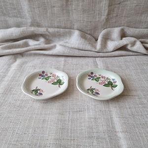 May include: Two small, off-white ceramic plates with a scalloped edge. Each plate features a floral design with pink flowers, green leaves, and purple berries. A light green border frames the design.