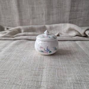 May include: A small, white ceramic jar with a lid, adorned with delicate floral patterns in shades of pink, blue, and green. The jar has a rounded shape and a small knob on top of the lid. The background is a neutral linen fabric.