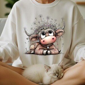 May include: A white sweatshirt featuring a cartoon illustration of a Highland cow with a flower crown and a bell around its neck. The cow is light brown and pink, with large eyes. A sleeping white cat rests in the foreground.