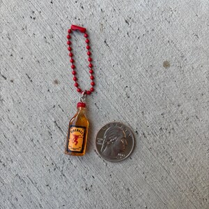 May include: Miniature Fireball bottle keychain with a red beaded chain. The bottle has a yellow liquid and a red cap, with the Fireball logo visible. A US quarter is next to the keychain for size comparison.