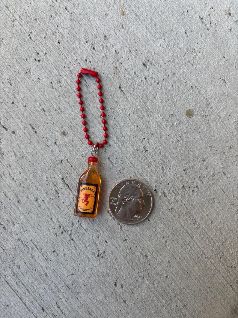 May include: Miniature Fireball bottle keychain with a red beaded chain. The bottle has a yellow liquid and a red cap, with the Fireball logo visible. A US quarter is next to the keychain for size comparison.