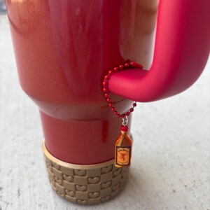 May include: A red insulated tumbler with a matching handle and a decorative charm. The charm is a miniature bottle of Fireball whiskey. The bottom of the tumbler has a tan, woven-like design.