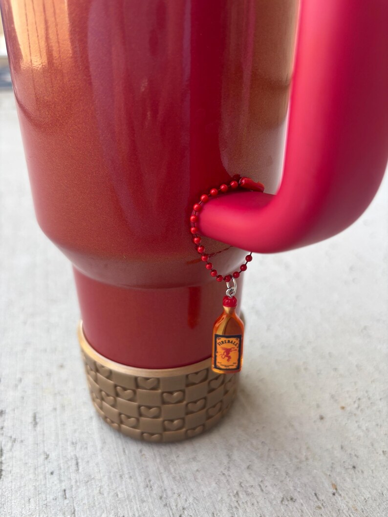 May include: A red insulated tumbler with a matching handle and a decorative charm. The charm is a miniature bottle of Fireball whiskey. The bottom of the tumbler has a tan, woven-like design.