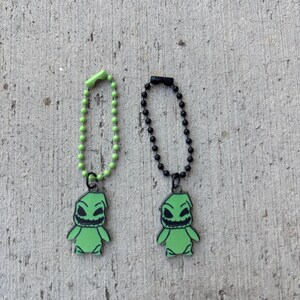 May include: Two green Oogie Boogie character charms with black outlines, each attached to a ball chain. One chain is green, the other black. The charms are on a textured gray surface.