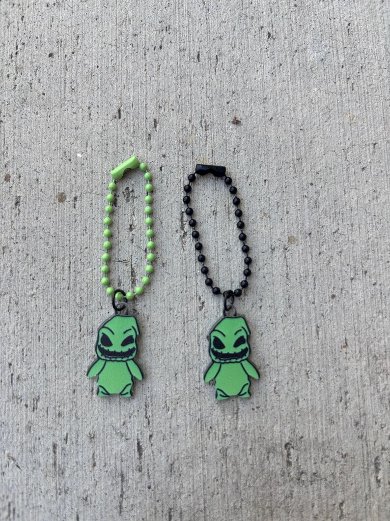May include: Two green Oogie Boogie character charms with black outlines, each attached to a ball chain. One chain is green, the other black. The charms are on a textured gray surface.