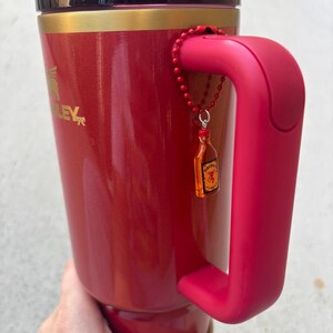 May include: A red Stanley travel mug with a matching handle and lid. The mug has a gold band near the top and a small charm of a liquor bottle attached to the handle. The mug is held by a person.