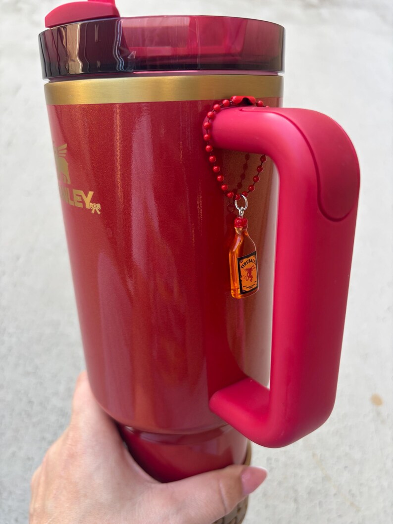May include: A red Stanley travel mug with a matching handle and lid. The mug has a gold band near the top and a small charm of a liquor bottle attached to the handle. The mug is held by a person.