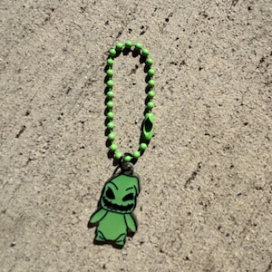 May include: A green charm featuring a cartoon character with a sinister grin, attached to a green ball chain. The charm is a vibrant green color with black accents, and the chain has a matching green clasp.