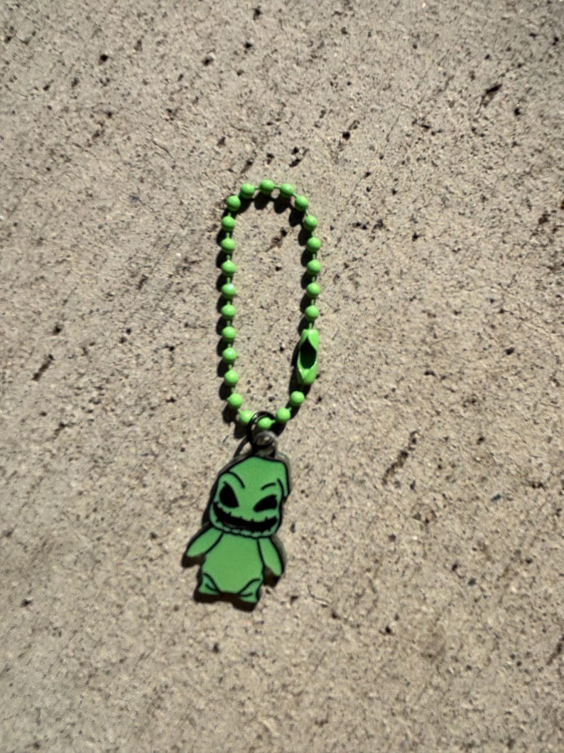 May include: A green charm featuring a cartoon character with a sinister grin, attached to a green ball chain. The charm is a vibrant green color with black accents, and the chain has a matching green clasp.
