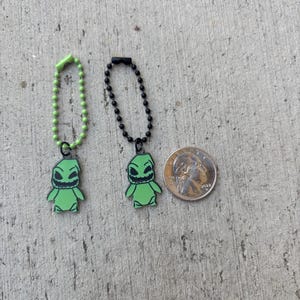 May include: Two green Oogie Boogie character charms with black outlines, each attached to a chain. One chain is green, the other black. A US quarter is next to the charms for size comparison.