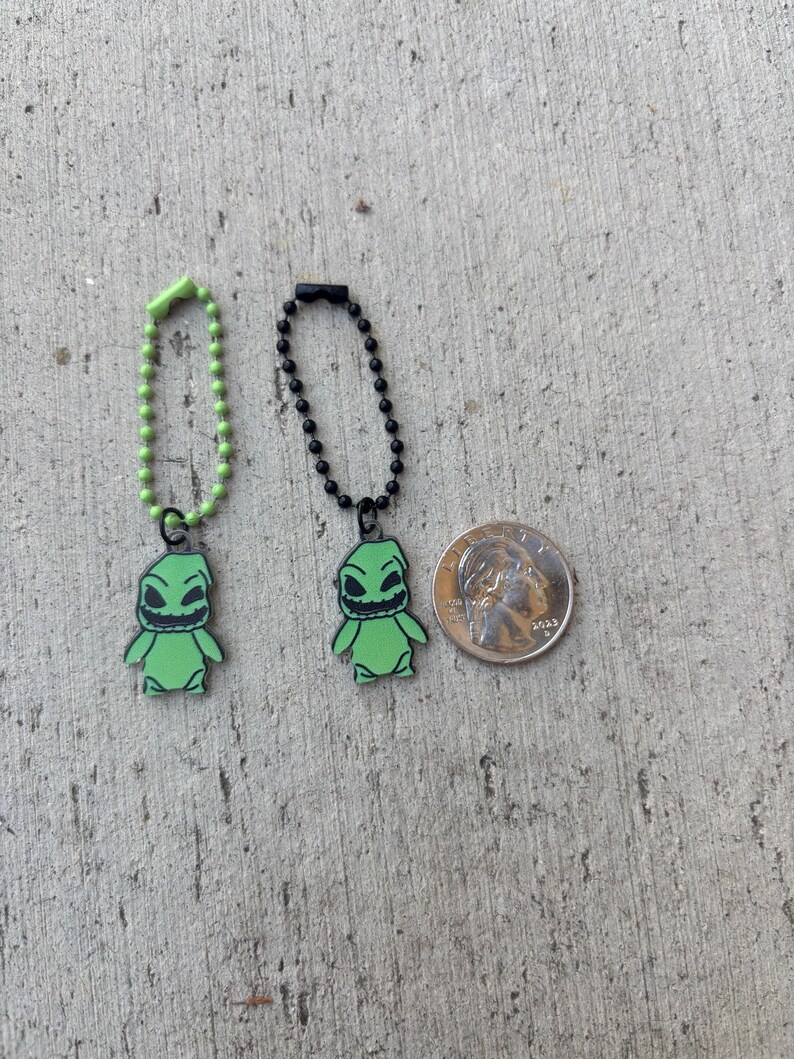 May include: Two green Oogie Boogie character charms with black outlines, each attached to a chain. One chain is green, the other black. A US quarter is next to the charms for size comparison.