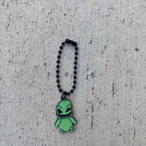 May include: A green Oogie Boogie charm on a black ball chain. The charm features a smiling Oogie Boogie character with black eyes and mouth. The charm is set against a textured gray background.