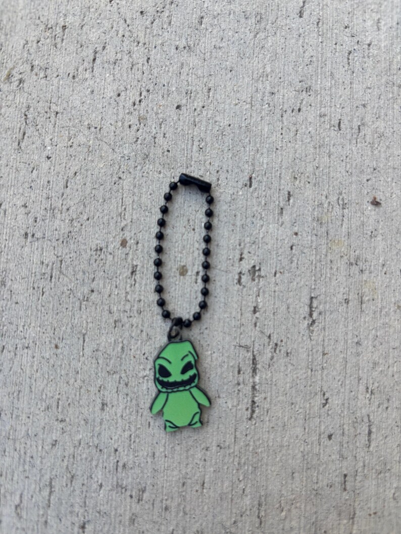May include: A green Oogie Boogie charm on a black ball chain. The charm features a smiling Oogie Boogie character with black eyes and mouth. The charm is set against a textured gray background.