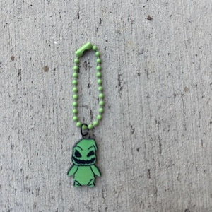 May include: A green keychain featuring a cartoon character with a sinister grin and black outlines. The character is attached to a green beaded chain with a matching clasp. The background is a textured gray surface.