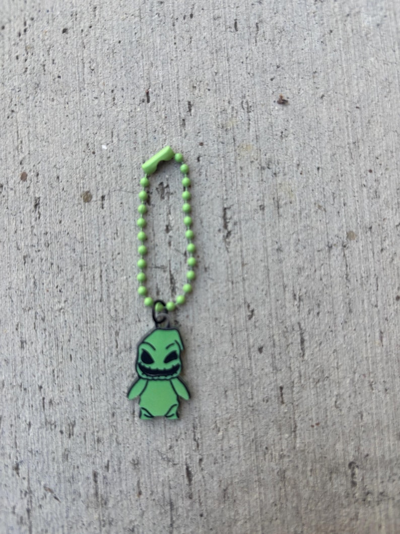 May include: A green keychain featuring a cartoon character with a sinister grin and black outlines. The character is attached to a green beaded chain with a matching clasp. The background is a textured gray surface.