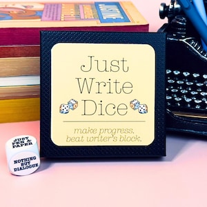 Peut inclure: Une boîte noire avec le texte "Just Write Dice" et des illustrations de dés. La boîte est à côté d'une pile de livres et d'une machine à écrire. Un petit dé avec le texte "Just Pen & Paper" est également visible.