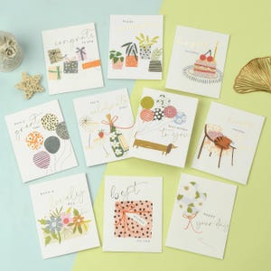 Puede incluir: Una colección de tarjetas de felicitación con varios diseños y mensajes. Las tarjetas presentan ilustraciones de regalos, plantas, globos, un perro salchicha, pastel y flores. Las letras doradas añaden un toque de elegancia, perfectas para celebraciones.