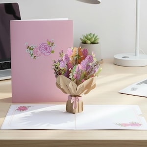 May include: A pink pop-up greeting card featuring a 3D paper flower bouquet. The bouquet includes pink, purple, and orange flowers, wrapped in brown paper and tied with a pink ribbon. A matching pink card is behind it.