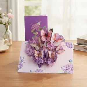 Op de afbeelding: Pop-up wenskaart met een 3D bloemstuk van paarse hortensia's en vlinders. De vlinders hebben roze en bruine vleugels. De kaart is open en onthult het ingewikkelde ontwerp, met een paarse kaart op de achtergrond.