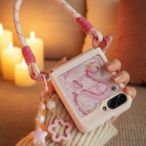 May include: A light pink phone case featuring a dragon illustration, a matching strap, and decorative charms. The case has a camera cutout and a hinged design. The strap is white with pink accents.